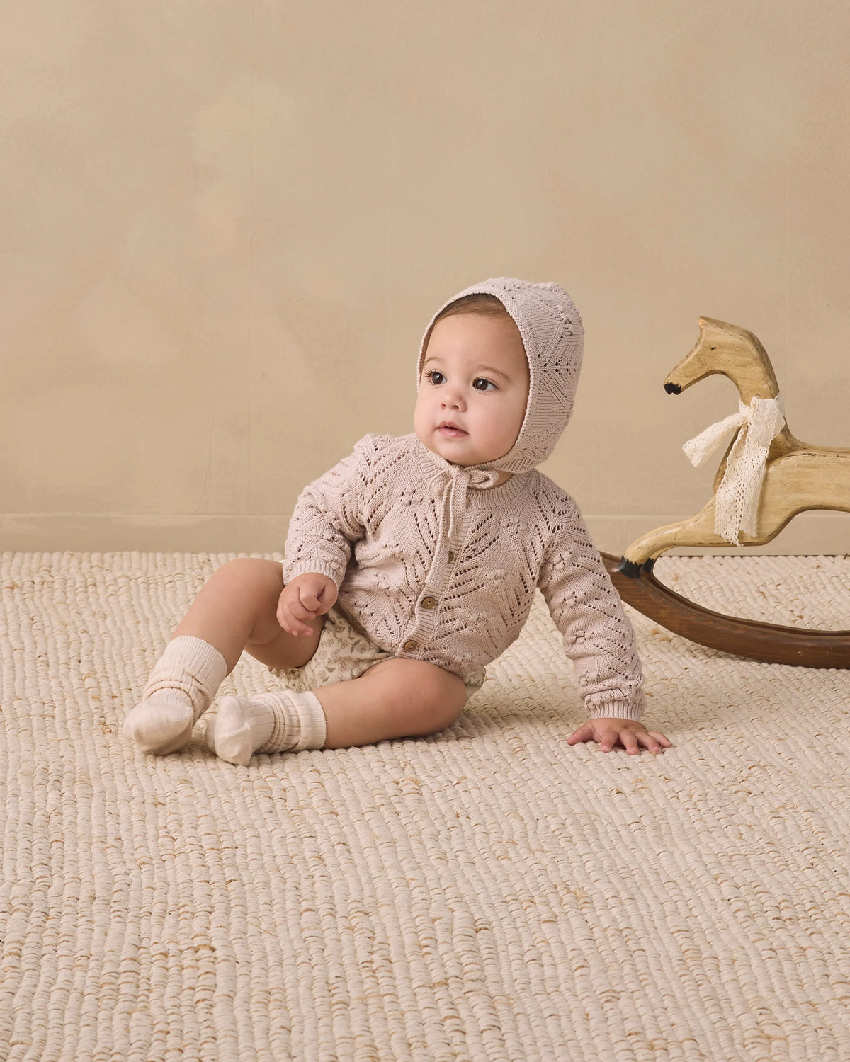 A baby wearing Quincy Mae’s Baby Girls’ Gathered Bloomer with elasticated leg openings sits on a textured rug next to a wooden rocking horse, paired with a knit hooded sweater and socks.