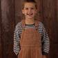 A young boy smiles while standing in front of a wooden wall, wearing the Rylee & Cru Baby Boys’ and Boys’ Relaxed Long Sleeve Tee in stripes with a ribbed neck and tan corduroy overalls featuring a chest pocket.
