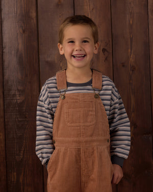 A young boy smiles while standing in front of a wooden wall, wearing the Rylee & Cru Baby Boys’ and Boys’ Relaxed Long Sleeve Tee in stripes with a ribbed neck and tan corduroy overalls featuring a chest pocket.
