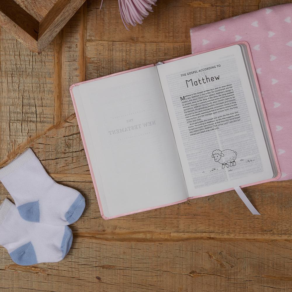 An open Baby's First KJV New Testament, Blue by Thomas Nelson is placed on a wooden surface alongside a pair of white baby socks and a pink blanket adorned with white hearts.