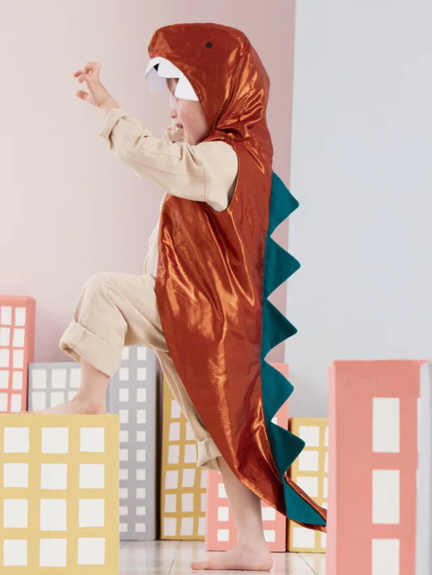 A child in a Meri Meri Dinosaur Costume, featuring spikes and a hooded jacket, appears to be playfully roaring, standing among cardboard cutout buildings.