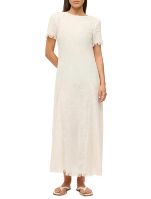 A woman wears the Staud Le Havre Dress, an off-white, short-sleeve linen dress with minimal design and frayed edges, paired with beige sandals, standing against a plain background.