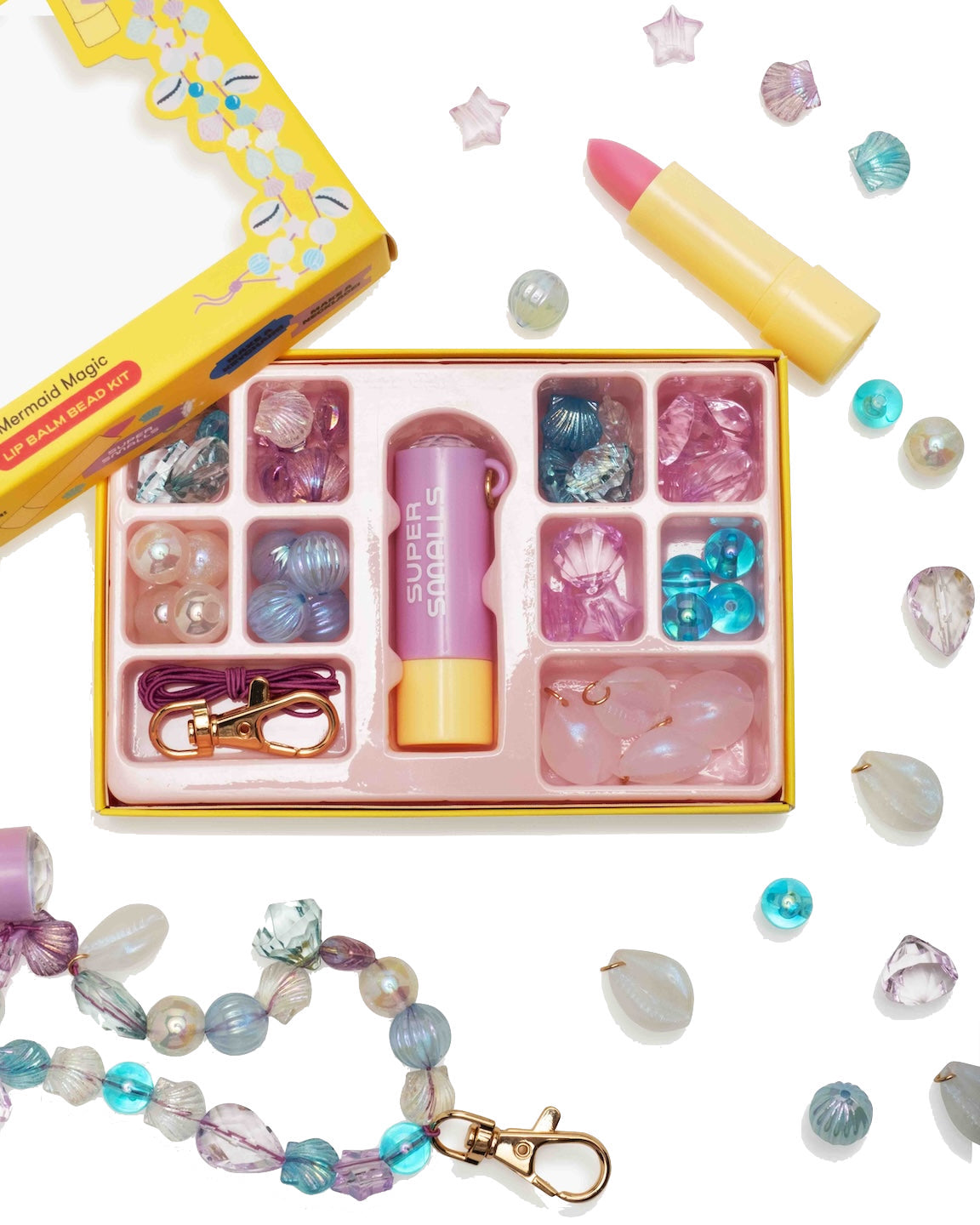 The Super Smalls Mermaid Magic Lip Balm Bead Kit includes beads, string, a clasp, a pink "Super Sparkle" flashlight, and lipstick-shaped Mermaid Magic Lip Balm, all arranged on a white surface.