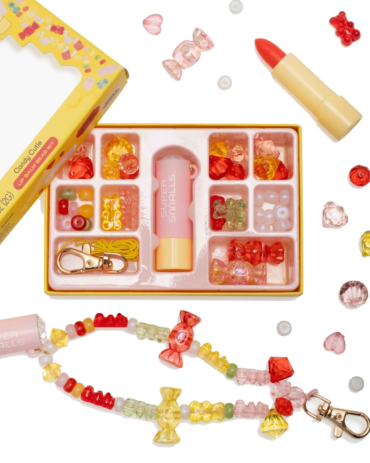 The Super Smalls Cutie Candy Lip Balm Bead Kit includes colorful beads, a strawberry-scented lip balm in a lipstick-shaped tube, yellow cord, and a finished bracelet—all arranged on a white background.