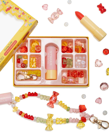 The Super Smalls Cutie Candy Lip Balm Bead Kit includes colorful beads, a strawberry-scented lip balm in a lipstick-shaped tube, yellow cord, and a finished bracelet—all arranged on a white background.