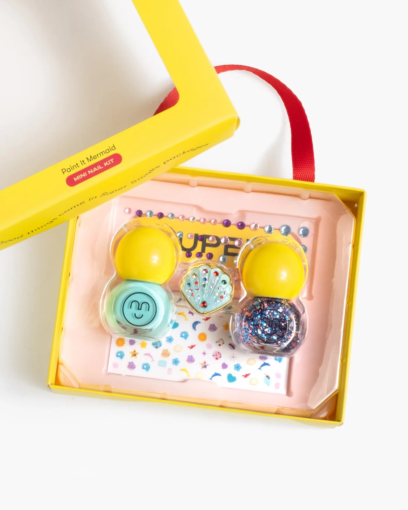 A yellow box holds the Super Smalls Paint It Mermaid Mini Nail Kit, featuring two yellow-capped bottles of peel-off nail polish, a seashell-shaped item, a blue container, and a sheet of shell nail stickers.