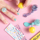 Someone uses the Super Smalls Paint It Mermaid Mini Nail Kit to apply blue and glitter peel-off polish, with shell nail stickers, gems, a nail file, and open polish bottles displayed on a pink surface.