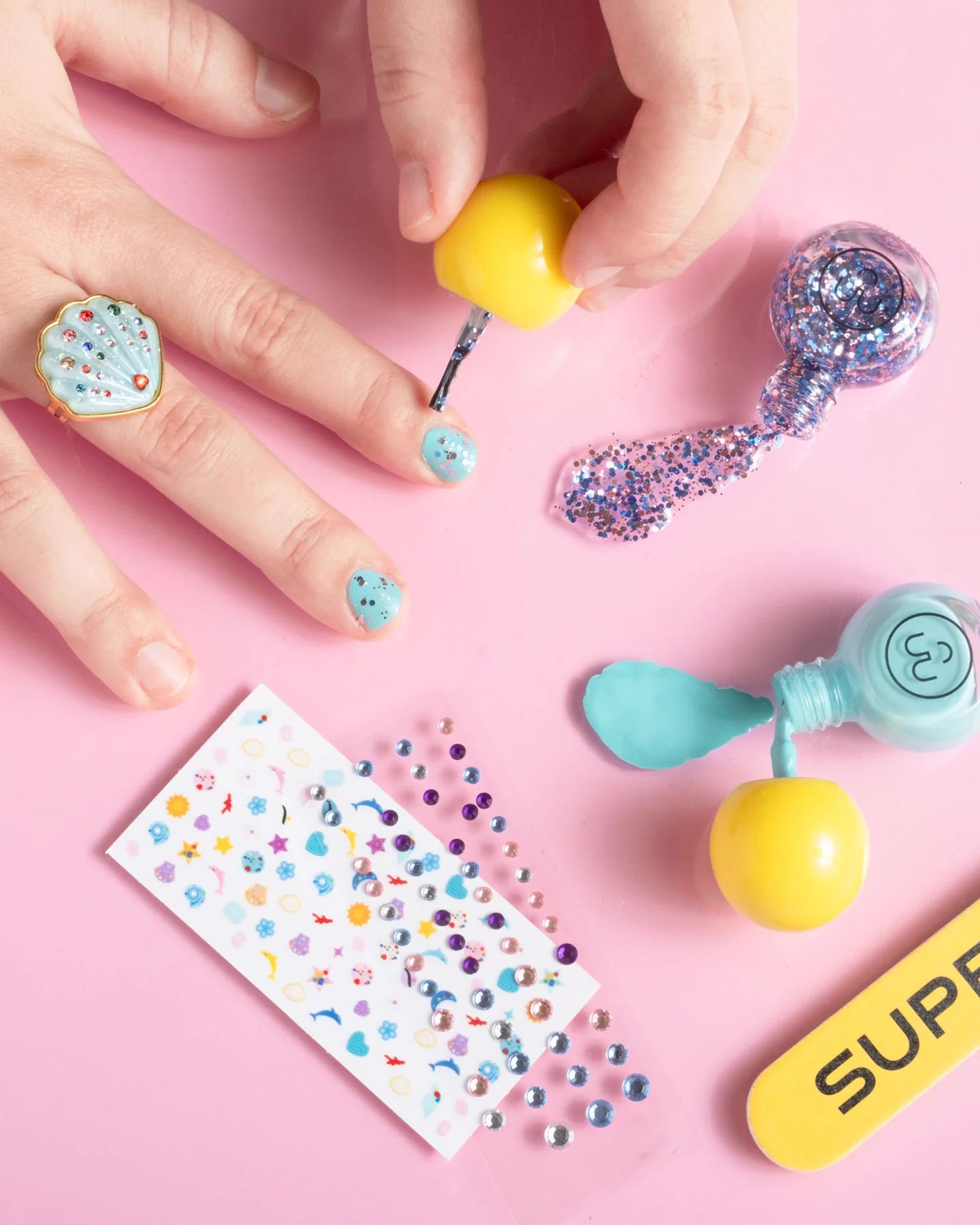 Someone uses the Super Smalls Paint It Mermaid Mini Nail Kit to apply blue and glitter peel-off polish, with shell nail stickers, gems, a nail file, and open polish bottles displayed on a pink surface.