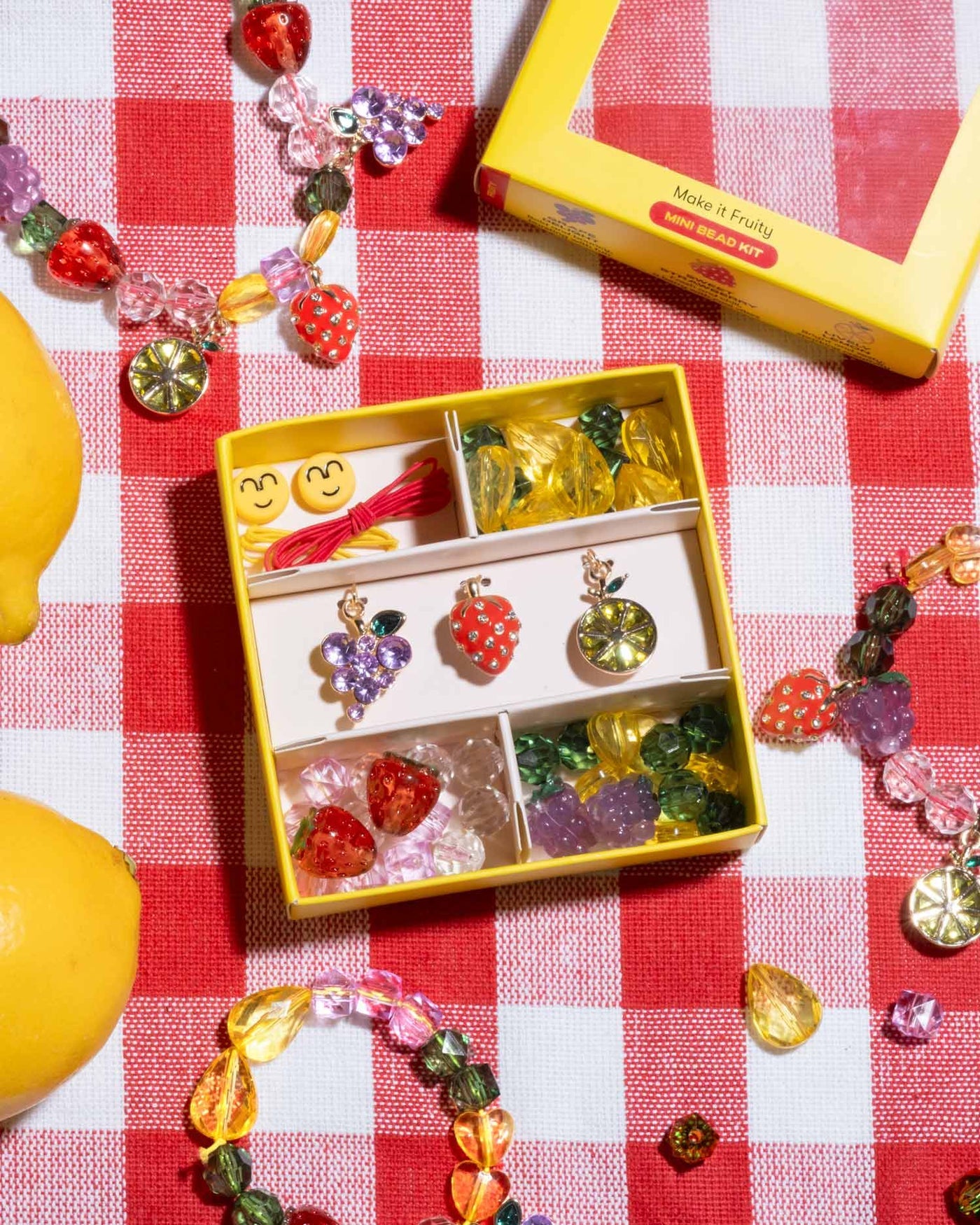 The Make It Fruity Mini Bead Kit by Super Smalls, featuring berry-bright beads, fruit charms, and string, is displayed on a red-and-white checkered surface next to lemons.