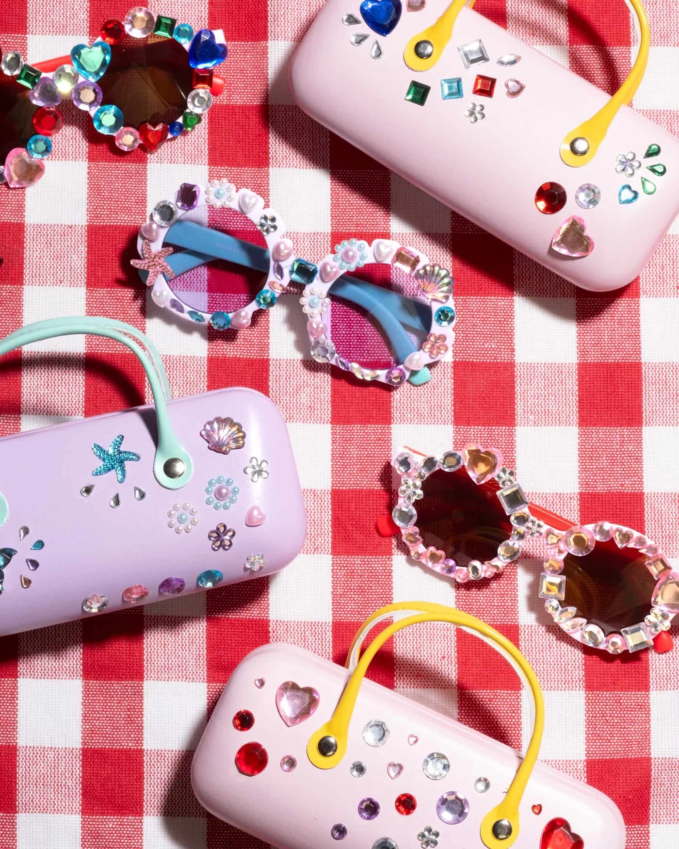 Super Smalls' Mermaid Splash & Sparkle Gemify Sunglasses & Case features decorative sunglasses and colorful cases with gems and stickers, arranged on a red-and-white checkered surface—ideal for kids’ crafting fun.