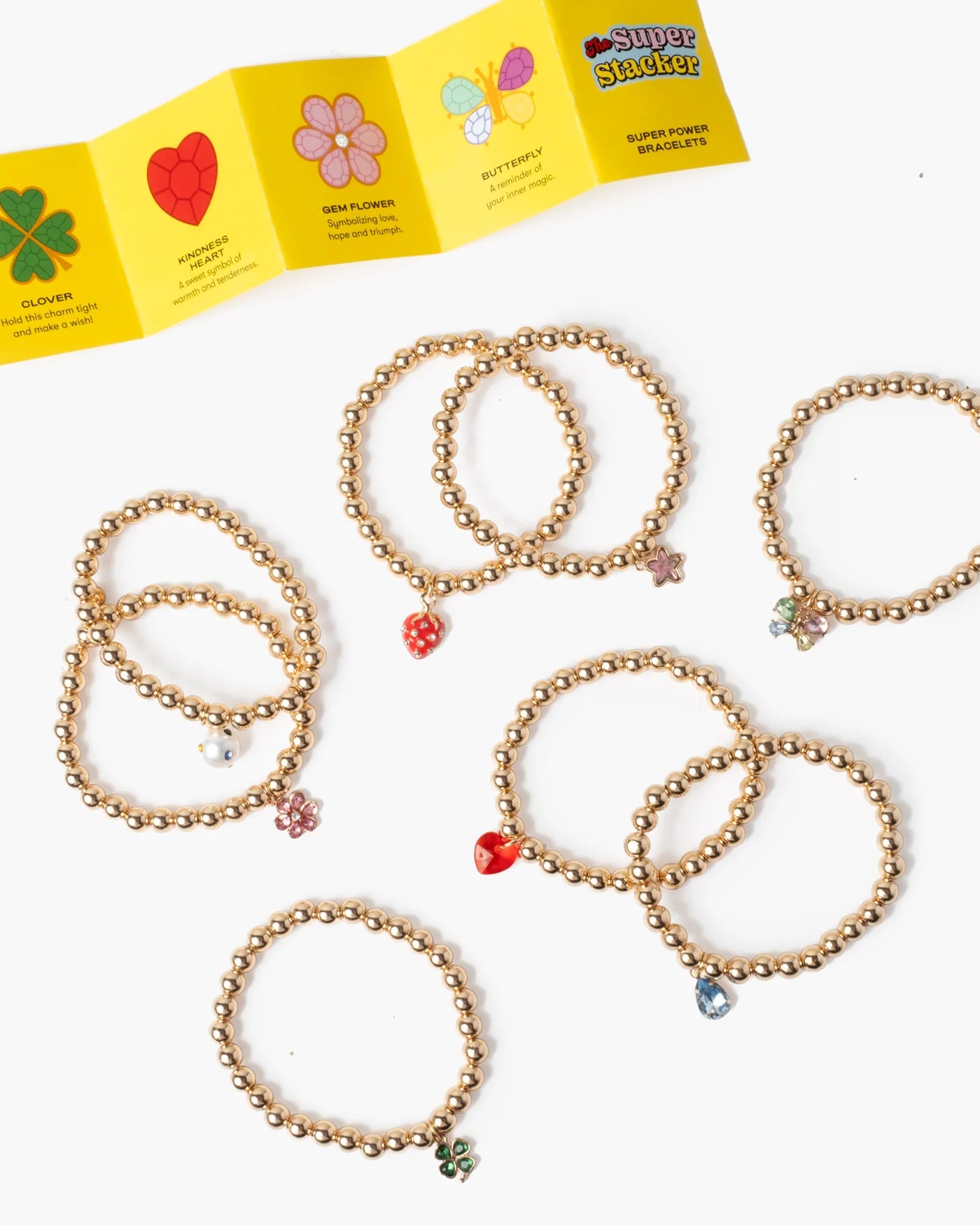 The Super Smalls Super Stack Bracelet Set includes seven gold beaded bracelets with colorful charms, arranged on a white surface beside a yellow folded instruction sheet—perfect for mixing and matching every day.
