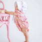 A baby girl in The Beaufort Bonnet Company's Pima Ramona Ruffle Bubble and a white bonnet stands holding a pink chair, gazing upward.