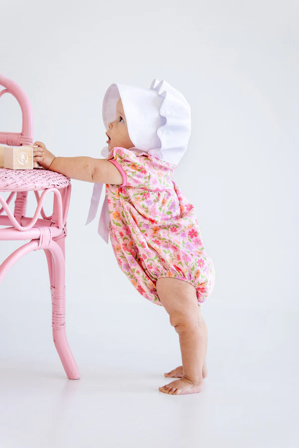 A baby girl in The Beaufort Bonnet Company's Pima Ramona Ruffle Bubble and a white bonnet stands holding a pink chair, gazing upward.