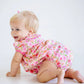 A blonde baby girl crawls on a white floor, smiling over her shoulder while wearing The Beaufort Bonnet Company's Baby Girls' Pima Ramona Ruffle Bubble.