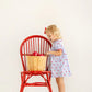 A young child in The Beaufort Bonnet Company’s Girls’ Mary Dal Dress stands by a red chair, reaching for apples in a basket—truly the apple of my eye.