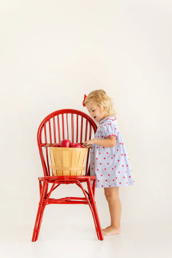 A young child in The Beaufort Bonnet Company’s Girls’ Mary Dal Dress stands by a red chair, reaching for apples in a basket—truly the apple of my eye.