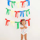 A young child stands before a white wall adorned with colorful ribbon garlands, wearing The Beaufort Bonnet Company's Baby Girls' Cindy Lou Bubble—a delightful piece for any little one's wardrobe.