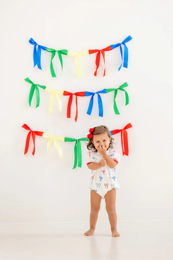 A young child stands before a white wall adorned with colorful ribbon garlands, wearing The Beaufort Bonnet Company's Baby Girls' Cindy Lou Bubble—a delightful piece for any little one's wardrobe.