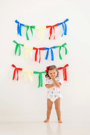 A young child stands before a white wall adorned with colorful ribbon garlands, wearing The Beaufort Bonnet Company's Baby Girls' Cindy Lou Bubble—a delightful piece for any little one's wardrobe.