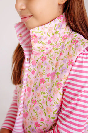 A girl wears The Beaufort Bonnet Company's Baby Girls' and Girl's Vaughn Vest in the Grand Cayman Gardens floral pattern over a pink and white striped shirt.