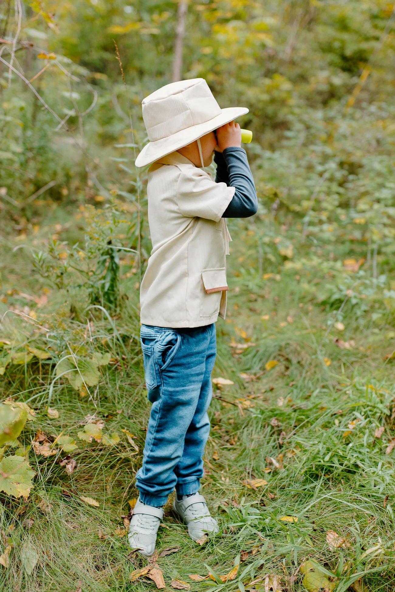 Dressed in the Great Pretenders Forest Guardian Set with Accessories, a child wearing a safari hat and casual clothes stands in grassy outdoors, holding binoculars and gazing into the distance—perfectly embodying the adventurous spirit of a young park ranger.