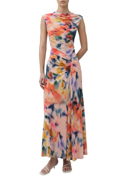 A woman wearing the Simkhai Acacia Jersey Midi Dress, featuring a sleeveless design, multicolored floral print, side ruching, and paired with white heeled sandals.