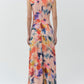 A person stands facing away, wearing the Simkhai Acacia Jersey Midi Dress—a sleeveless, draped design featuring a colorful abstract pattern—paired with low heels against a plain light background.