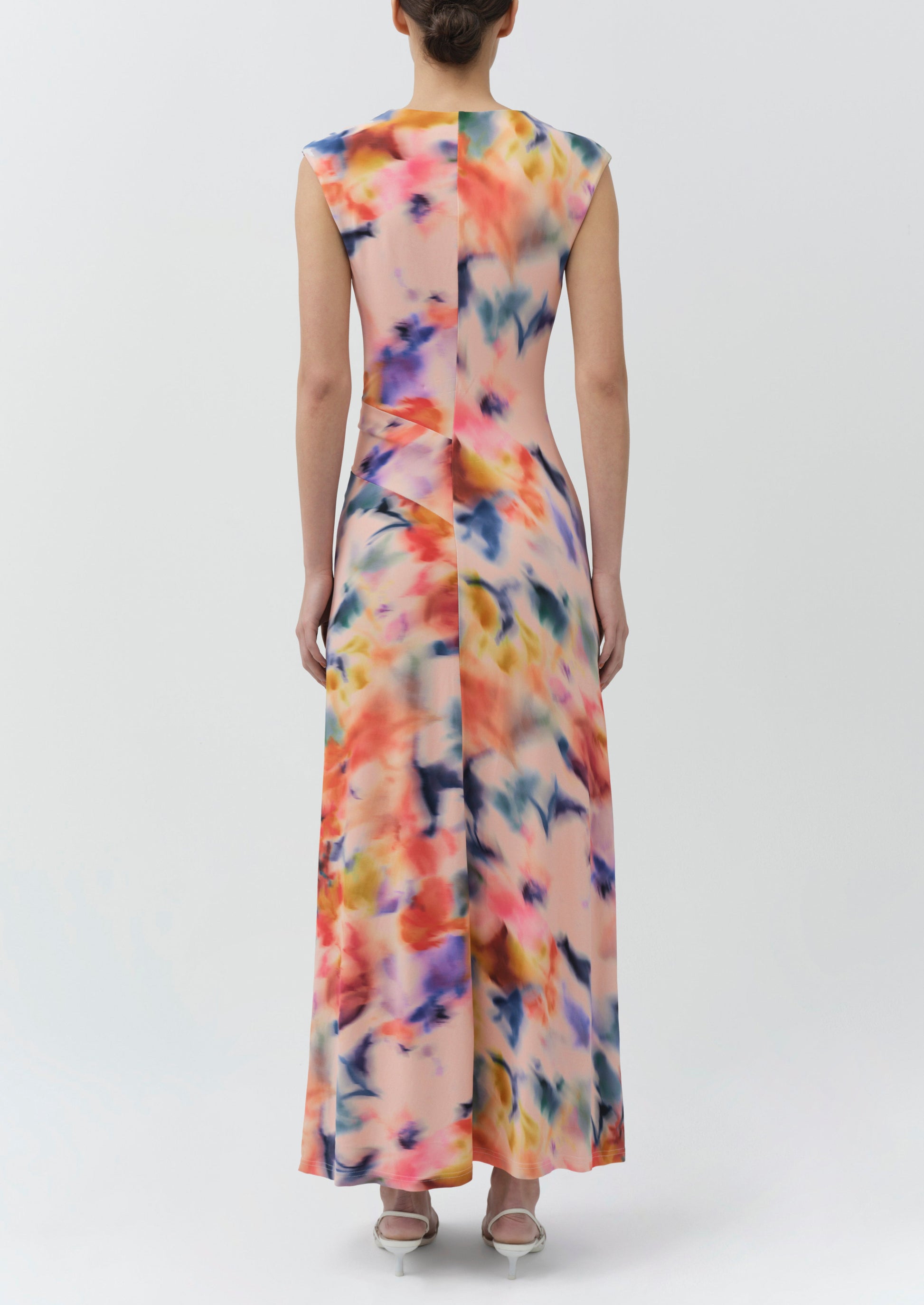 A person stands facing away, wearing the Simkhai Acacia Jersey Midi Dress—a sleeveless, draped design featuring a colorful abstract pattern—paired with low heels against a plain light background.