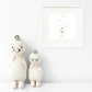 Two Harlow the Swan plush toys by Cuddle + Kind stand by a white wall, crowned and elegant; above them is a framed print that says, "With brave wings she flies," inspiring thoughts of courage.
