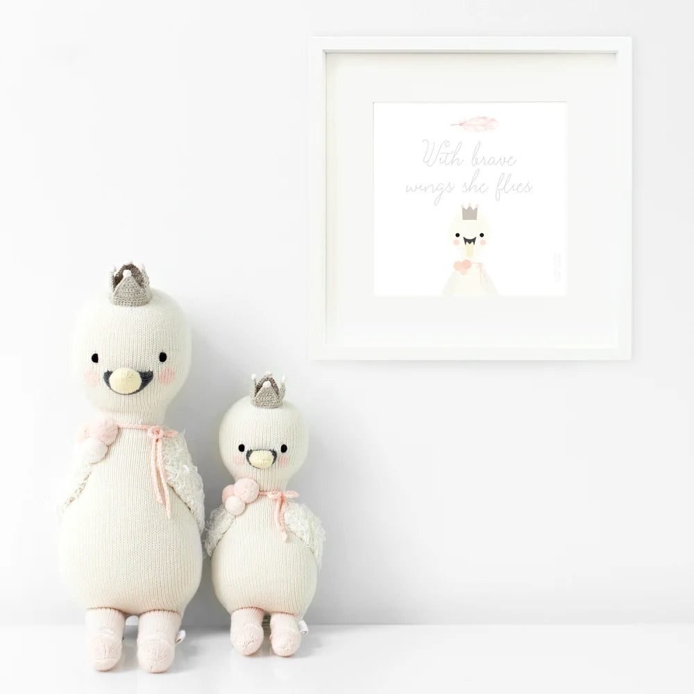 Two Harlow the Swan plush toys by Cuddle + Kind stand by a white wall, crowned and elegant; above them is a framed print that says, "With brave wings she flies," inspiring thoughts of courage.