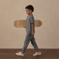 A young boy wears the Rylee & Cru Boys’ Cove Essential Pocket Tee as he walks indoors holding a skateboard under his arm, set against a beige background with a wood floor.