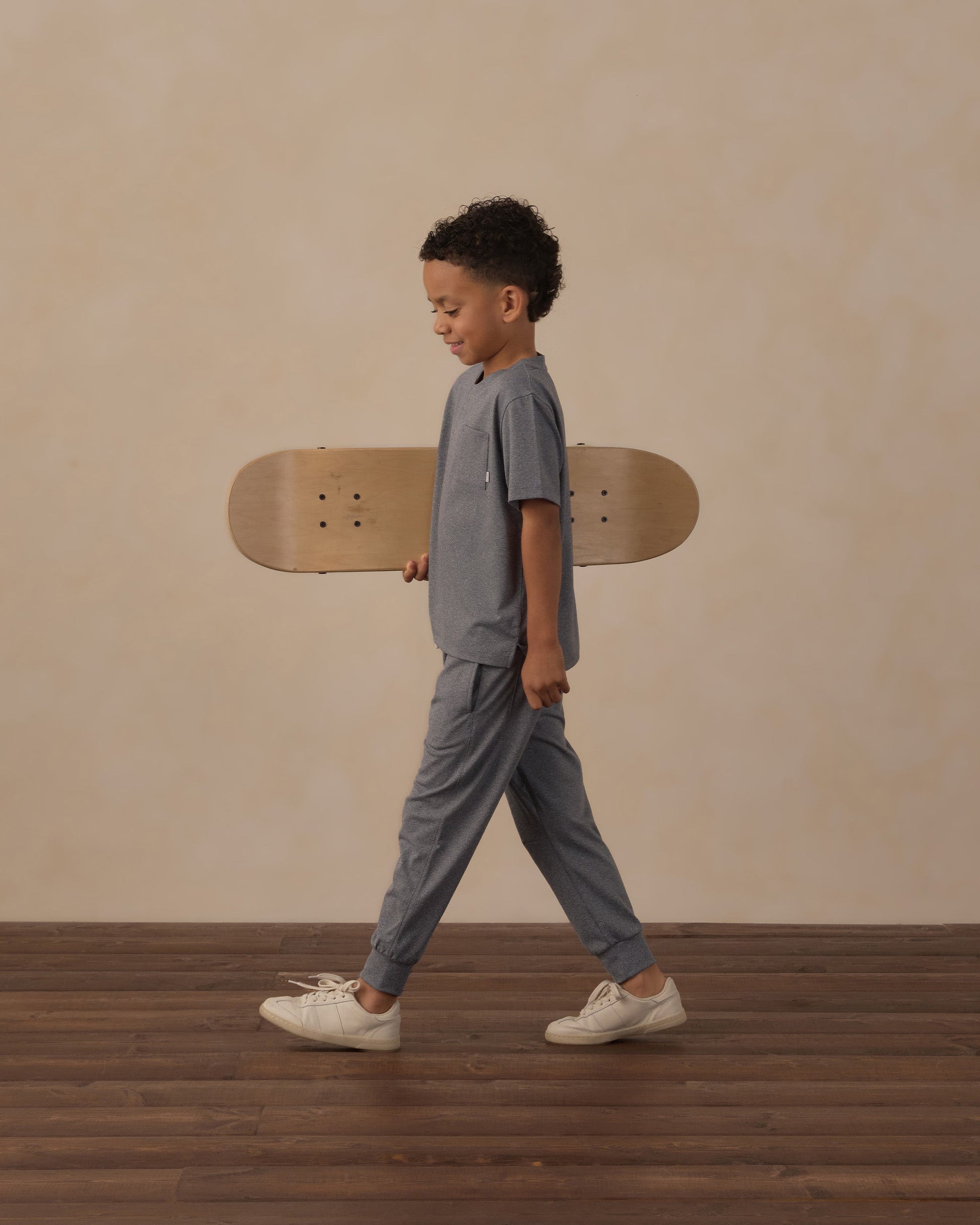 A young boy wears the Rylee & Cru Boys’ Cove Essential Pocket Tee as he walks indoors holding a skateboard under his arm, set against a beige background with a wood floor.