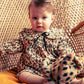 A baby in a Louis Louise Baby Girls' Benjamina Corduroy Leopard Dress sits on a wicker chair, holding a matching leopard print stuffed animal.