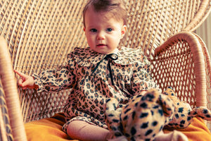 A baby in a Louis Louise Baby Girls' Benjamina Corduroy Leopard Dress sits on a wicker chair, holding a matching leopard print stuffed animal.