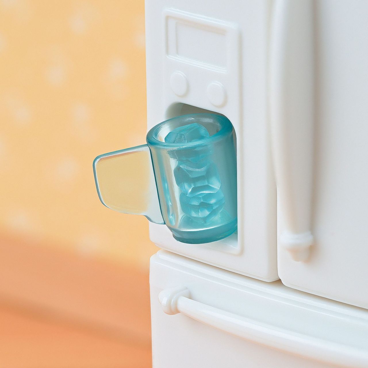 A blue cup filled with ice cubes sits in the dispenser of a white Playful Starter Furniture Set refrigerator from Calico Critters, inviting imaginative play against a peach-colored background.