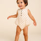 A toddler in the Lou Lou & Company Baby Cloud Muslin Bubble Romper, featuring a beige checkered pattern with buttons and convenient bottom snaps, stands barefoot on a light background, smiling and looking to the side—a true warm-weather essential.