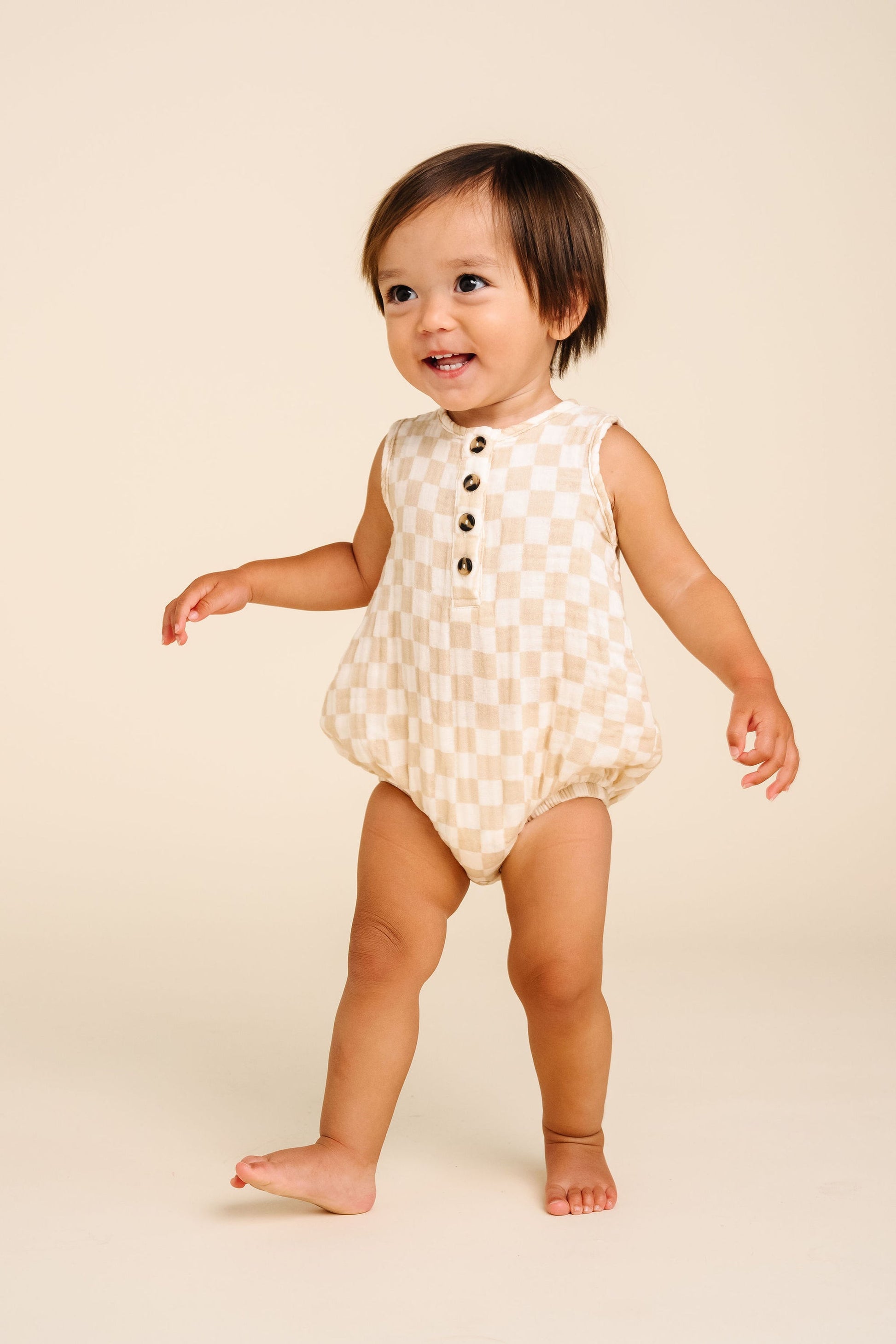 A toddler in the Lou Lou & Company Baby Cloud Muslin Bubble Romper, featuring a beige checkered pattern with buttons and convenient bottom snaps, stands barefoot on a light background, smiling and looking to the side—a true warm-weather essential.