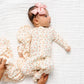 A baby girl dressed in the Lou Lou & Company Baby Girls’ Lottie Zipper Footie lies on a white blanket, a pink bow atop her head, while an adult hand gently supports her side—highlighting the cozy charm of these comfy pajamas designed for peaceful sleep.