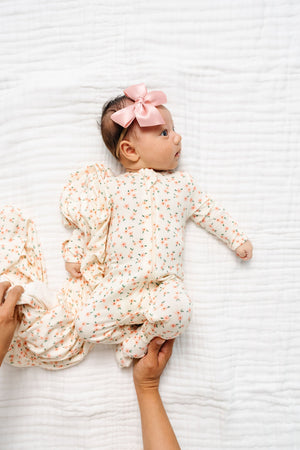 A baby girl dressed in the Lou Lou & Company Baby Girls’ Lottie Zipper Footie lies on a white blanket, a pink bow atop her head, while an adult hand gently supports her side—highlighting the cozy charm of these comfy pajamas designed for peaceful sleep.