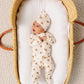 A baby wearing the Lou Lou & Company Baby Teddy Zipper Footie and a matching hat with bear faces lies in a woven basket on a white padded mattress.