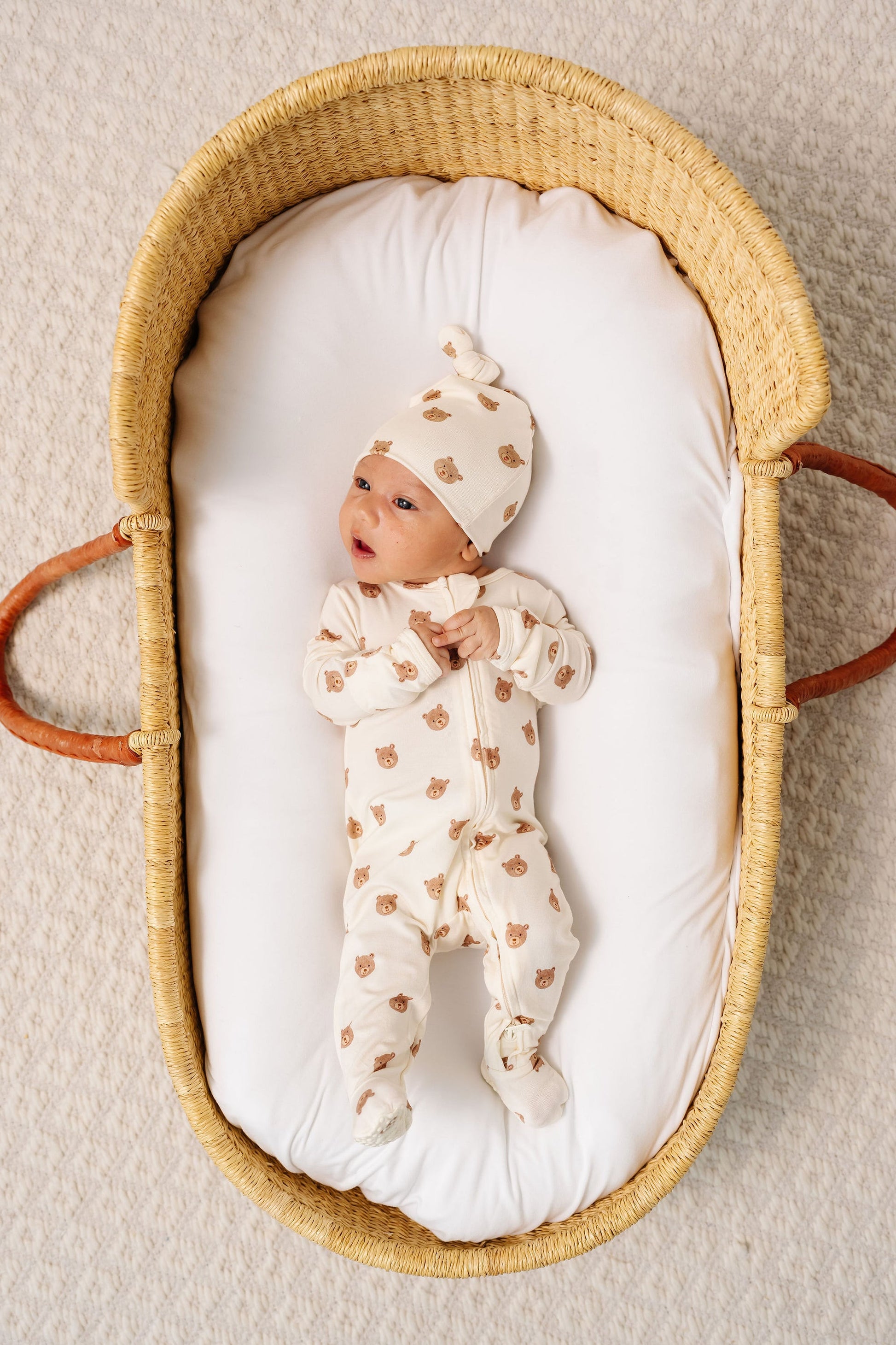 A baby wearing the Lou Lou & Company Baby Teddy Zipper Footie and a matching hat with bear faces lies in a woven basket on a white padded mattress.