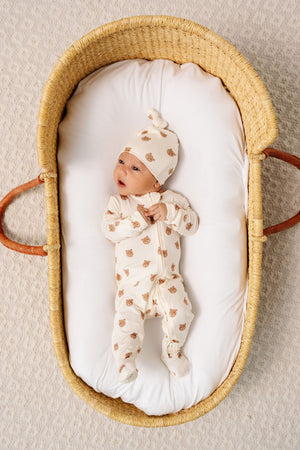 A baby wearing the Lou Lou & Company Baby Teddy Zipper Footie and a matching hat with bear faces lies in a woven basket on a white padded mattress.