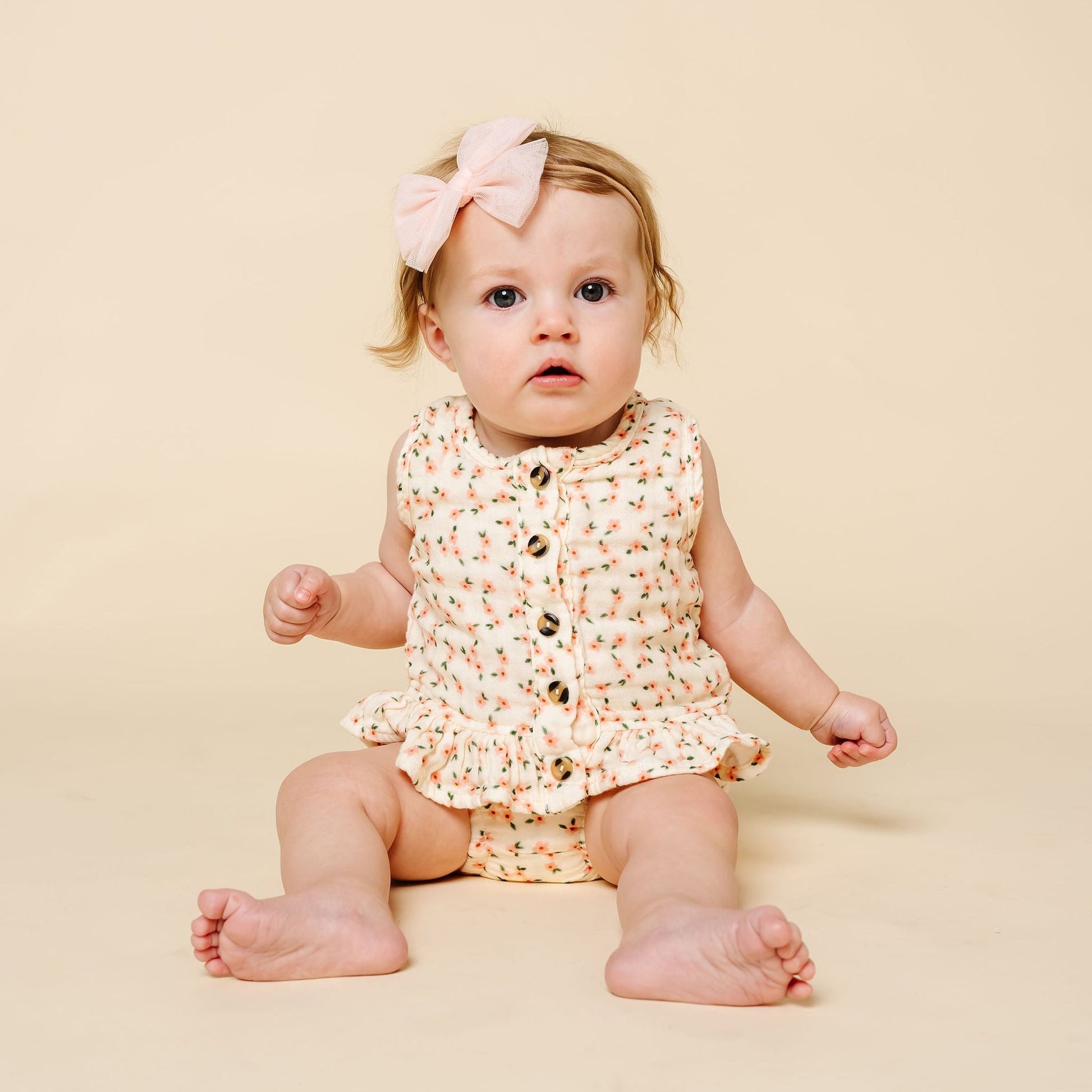 A baby wears the Lou Lou & Company Baby Girls’ Ruffle Tank & Bloomer Set, featuring a sleeveless, floral-patterned ruffle tank made from breathable cotton and a large pink bow headband, while sitting on a beige background and looking ahead.