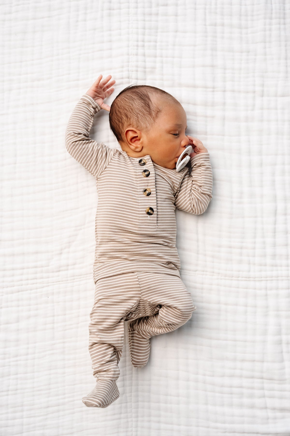 A baby wearing the Lou Lou & Company Baby Boys’ and Boys’ Banks Top & Bottoms sleeps on a white quilted blanket, holding a pacifier near their mouth.