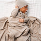 A baby in the Lou Lou & Company Baby Boys’ Banks Knotted Gown, complete with a matching hat and coordinated blanket for easy diaper changes, sleeps peacefully on a white textured surface while holding a pacifier.