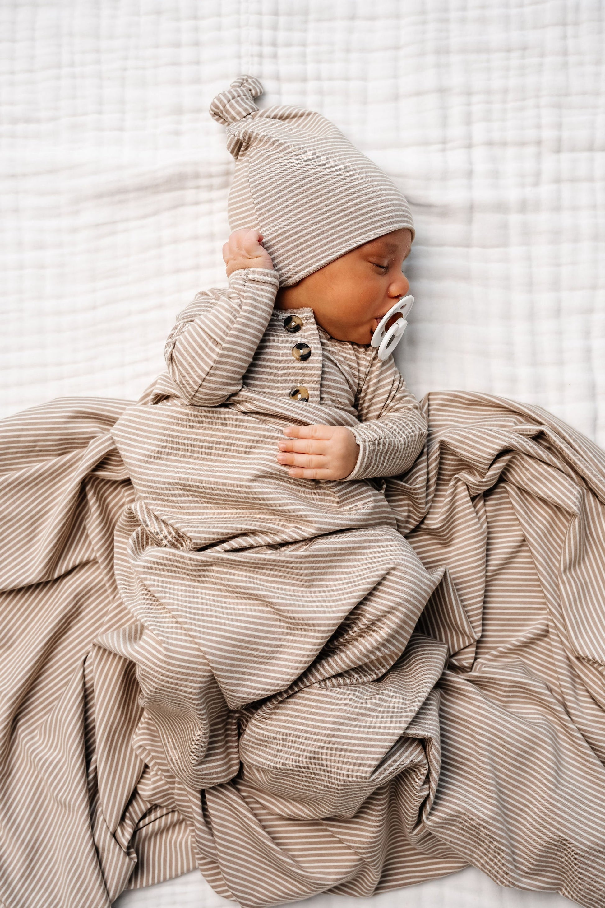 A baby in the Lou Lou & Company Baby Boys’ Banks Knotted Gown, complete with a matching hat and coordinated blanket for easy diaper changes, sleeps peacefully on a white textured surface while holding a pacifier.