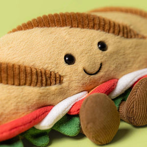 The Jellycat Amuseables Caprese Baguette is a plush toy shaped like a smiling sandwich, featuring brown bread, green lettuce, and colorful fillings, shown on a light green background.