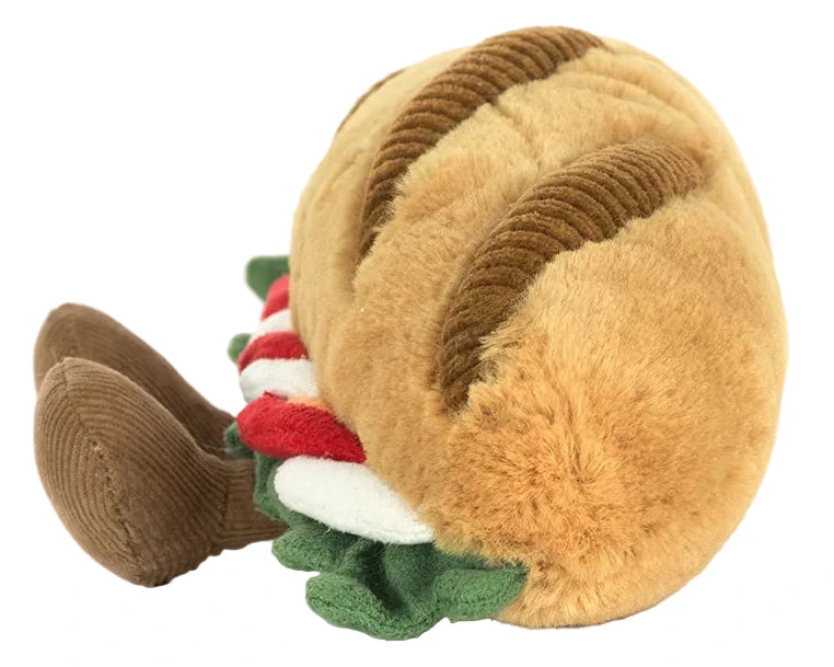 The Jellycat Amuseables Caprese Baguette is a plush toy shaped like a sandwich, featuring a brown bun, green lettuce, red tomato, and two adorable brown legs.
