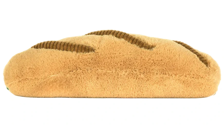 The Jellycat Amuseables Caprese Baguette is a plush toy shaped like a loaf of bread with a light brown color and dark brown textured lines on top.