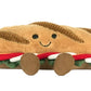 The Jellycat Amuseables Caprese Baguette is a plush toy sandwich with a smiling face, brown bread, green lettuce, red and white layers, and two cute brown feet.